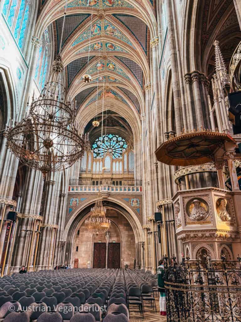 Das Innere der Votivkirche mit seinen hohen Kreuzrippengewölben, die mit farbenfrohen Fresken und kunstvollen Kronleuchtern geschmückt sind. Stuhlreihen säumen das Kirchenschiff und führen zu der großen Orgel und einer verzierten Kanzel mit vergoldeten Reliefs, die die reichen architektonischen Details der Kirche hervorheben.