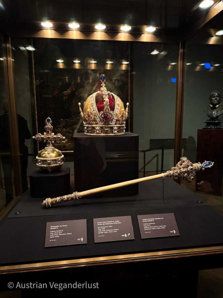 Eine Vitrine mit der Kaiserkrone des Heiligen Römischen Reiches, dem Reichsapfel und dem Zepter, ausgestellt in der Wiener Schatzkammer. Die Krone ist mit Gold, Perlen und Edelsteinen verziert, während der Reichsapfel und das Zepter kunstvoll gestaltet sind. Drei Informationstafeln mit Beschreibungen der Objekte befinden sich davor.