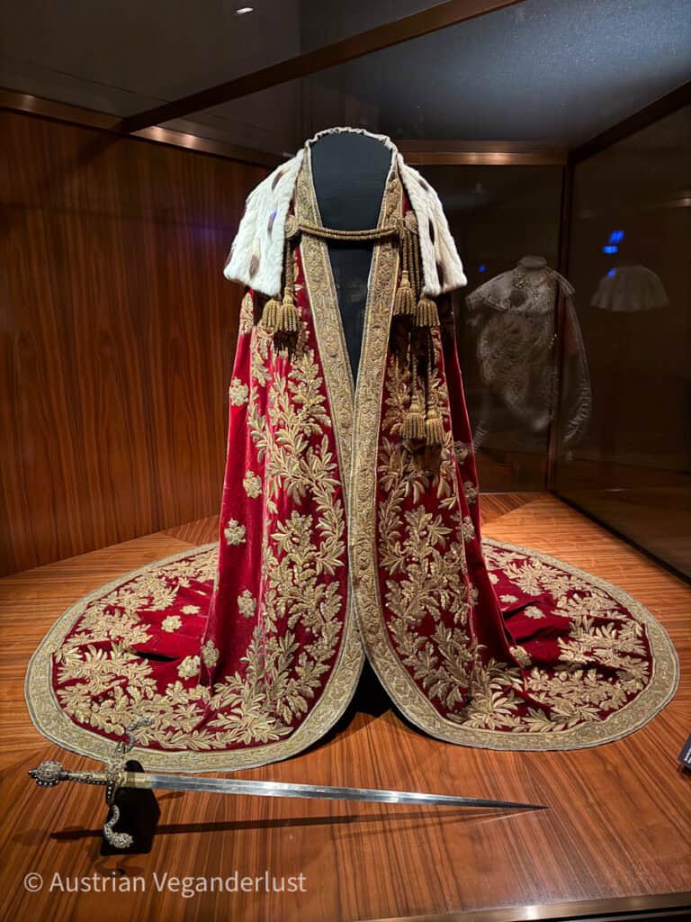 Ein prächtiger, roter Krönungsmantel mit goldenen Stickereien und Hermelinbesatz, ausgestellt in einer Vitrine in der Wiener Schatzkammer. Der Mantel hat ein aufwendiges Muster mit Blattmotiven und Quastenverzierungen. Davor liegt ein Schwert mit einem kunstvoll verzierten Griff.