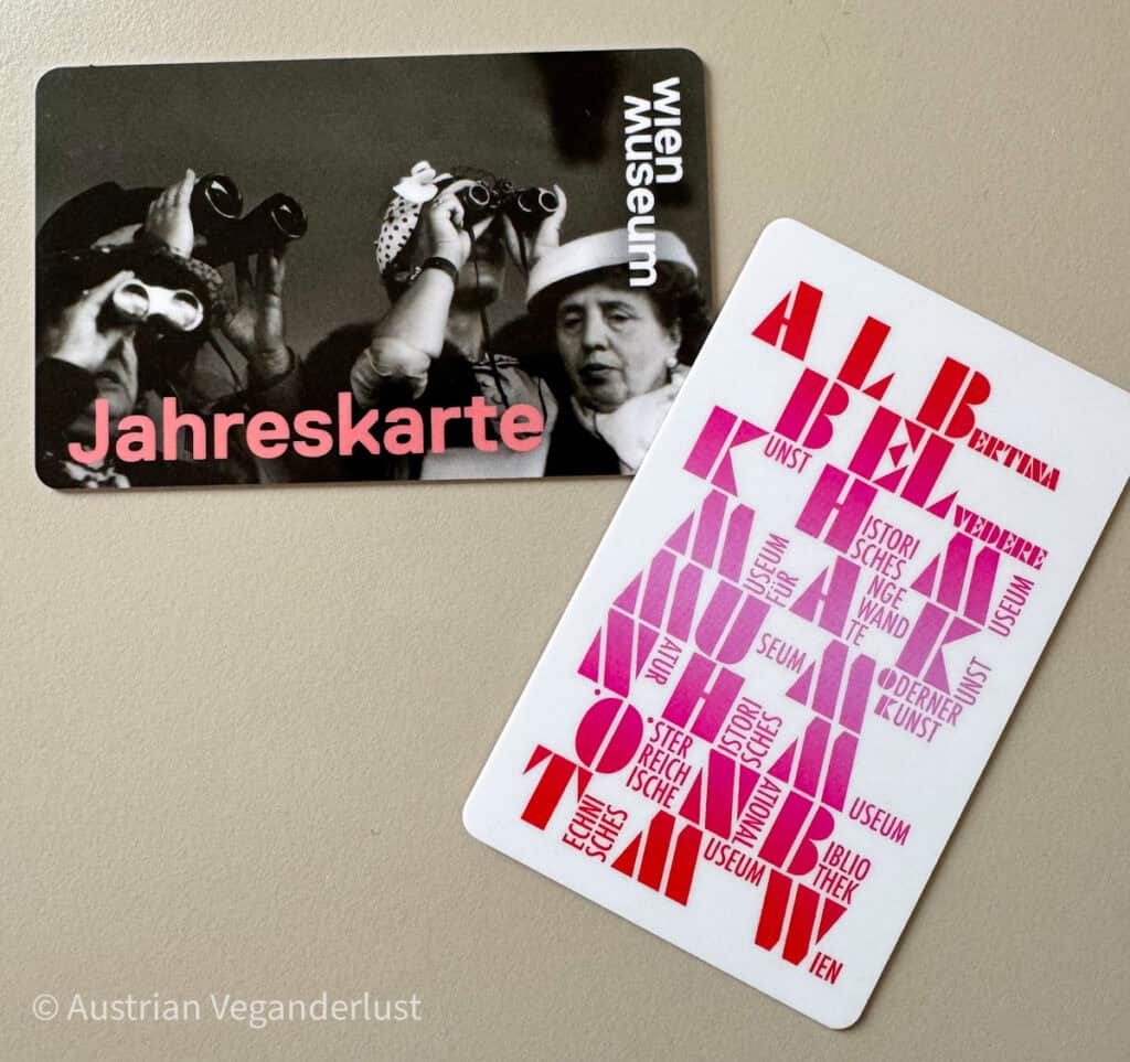 Zwei Museumskarten liegen auf einer beige Tischoberfläche. Die linke Karte ist eine Jahreskarte für das Wien Museum, mit einem historischen Schwarz-Weiß-Foto von Frauen, die durch Ferngläser schauen, und dem Wort „Jahreskarte“ in roter Schrift. Die rechte Karte zeigt eine moderne Typografie mit in Rot und Pink gesetzten Wörtern wie „ALBERTINA“, „MUSEUM“, „WIEN“ und „KUNST“.