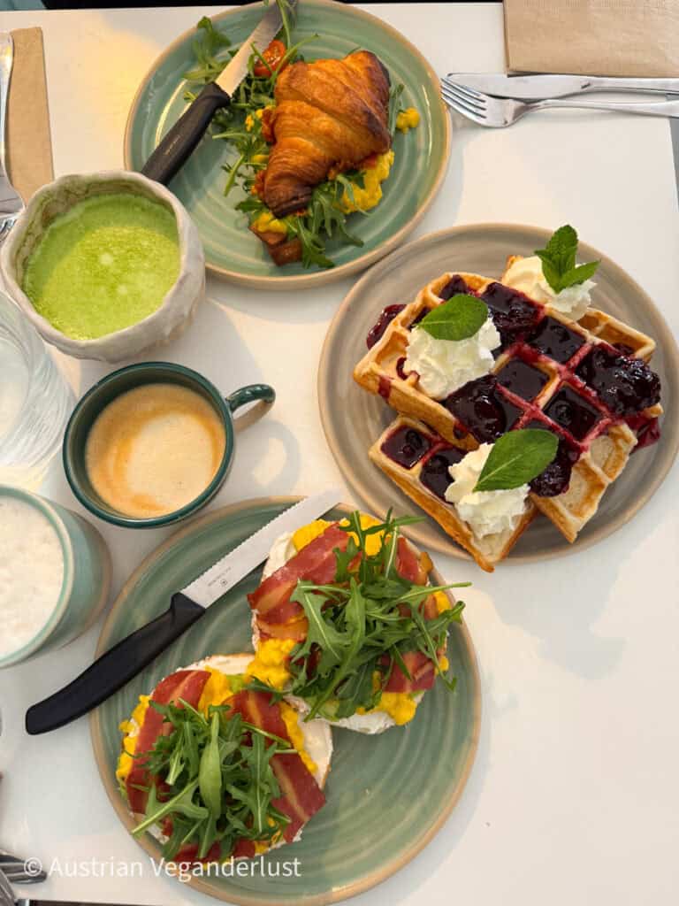 Ein lebendiger veganer Brunch mit knusprigen Waffeln mit Schlagsahne und Beerenkompott, einem Croissant-Sandwich mit Rucola und Tofu-Rührei, Matcha-Latte und offenen Bagels mit Frischkäse, Tofu-Rührei, veganem Speck und Rucola.