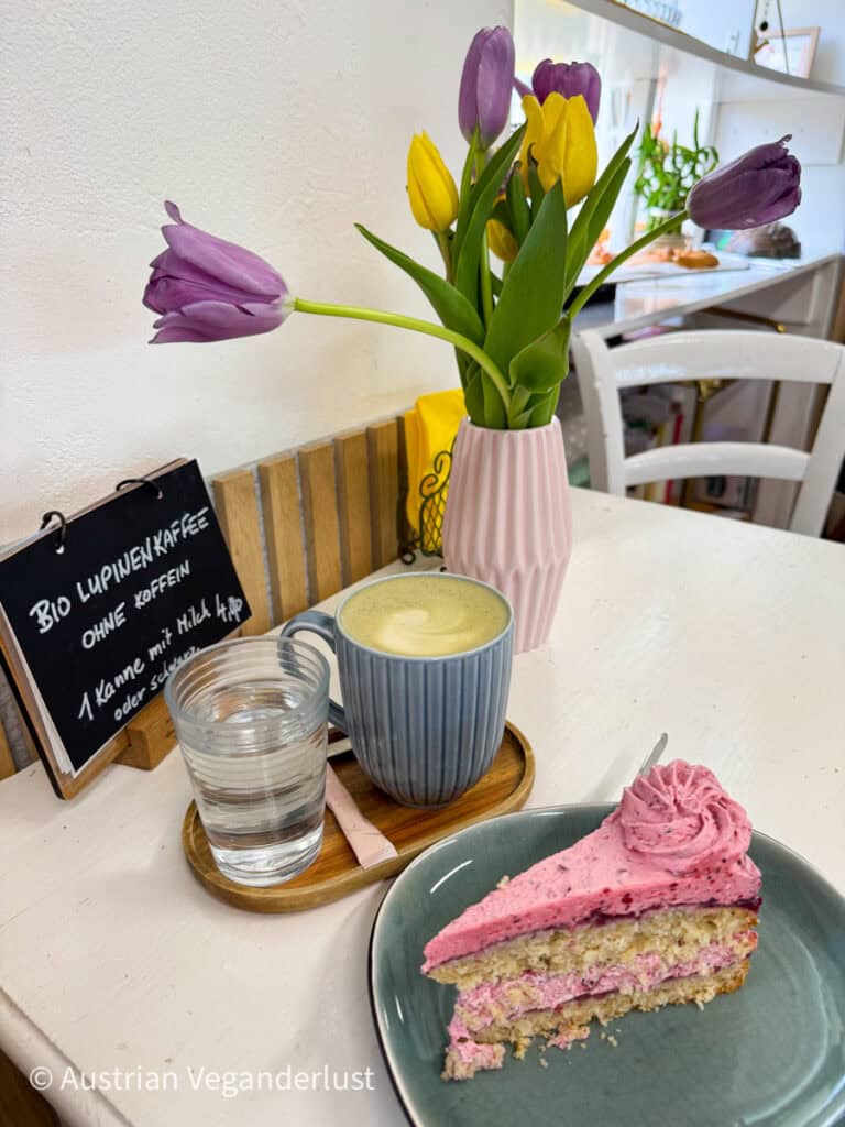Ein gemütlicher Cafétisch mit einem Stück rosa Schichtkuchen, einer blauen Tasse Lupinenkaffee und einem Glas Wasser, eingerahmt von einer Vase mit frischen lila und gelben Tulpen. Auf einer kleinen Kreidetafel steht "Bio Lupinenkaffee ohne Koffein - 1 Kanne mit Milch & Sirup oder Schwarz."