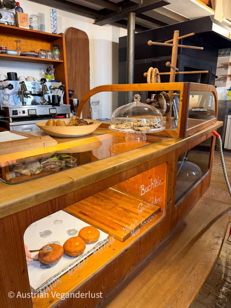 Eine gemütliche Bäckertheke aus Holz mit einer Glasvitrine, in der Gebäck präsentiert wird. Handgeschriebene Etiketten auf dem Glas weisen auf verschiedene Leckereien hin, darunter "Buchteln" und "Nougat-Marzipan". Ein Mitarbeiter greift mit einer Zange nach frisch gebackenen Krapfen, während Kekse und Bagels auf Holzständern ausgestellt sind. Hinter der Theke stehen eine Kaffeemaschine und Regale mit Bechern und Zutaten, die die einladende Atmosphäre abrunden.