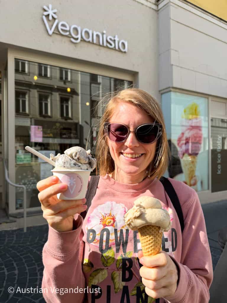 Tina mit Sonnenbrille und "Powered by Plants"-Sweatshirt hält an einem sonnigen Tag vor dem Veganista-Laden zwei vegane Eissorten - eine Kugel in einer Waffeltüte und eine andere in einem kleinen Pappbecher. Die Schaufensterfront hinter ihr zeigt das Veganista-Logo und ein leuchtendes Eiscreme-Poster im Schaufenster.