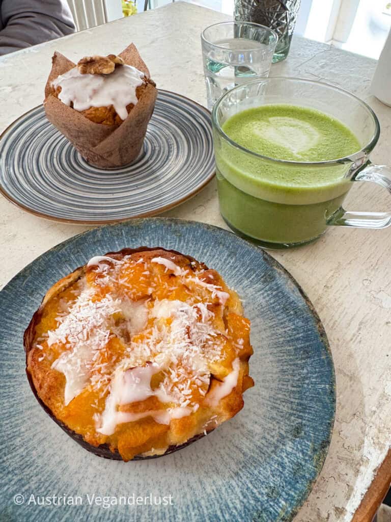 Ein heller und farbenfroher Cafétisch mit einem saftigen orangefarbenen Kokosnusskuchen mit Zuckerguss, einem Walnussmuffin mit Glasur, einem Glas Matcha Latte und einem Glas Wasser in sanftem Tageslicht.