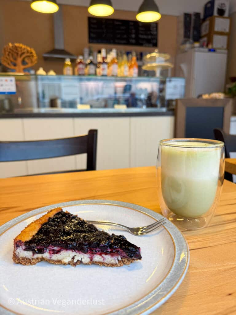 Ein Stück Beerenkäsekuchen mit bröckeliger Kruste und einem glänzenden, dunklen Fruchtbelag auf einem Keramikteller, daneben ein cremiger Matcha Latte in einem doppelwandigen Glas. Im Hintergrund ist eine gemütliche Café-Theke mit Flaschen, Gebäck und einer warmen Atmosphäre zu sehen.