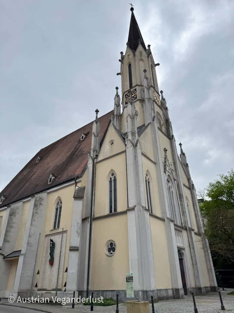 Das Äußere der Pfarrkirche Melk, ein cremefarbenes Gebäude im gotischen Stil mit spitzen Bögen, einem hohen Turm und kunstvollem Mauerwerk vor einem bewölkten Himmel.