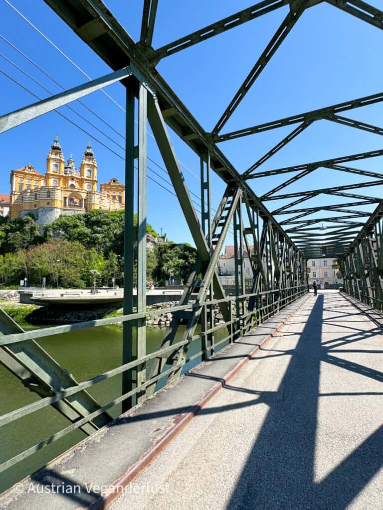 Durch die Struktur einer alten grünen Eisenbrücke blickst du auf das Stift Melk, das im Sonnenlicht glänzt und dessen starke Schatten ein dramatisches Muster auf dem Gehweg bilden.