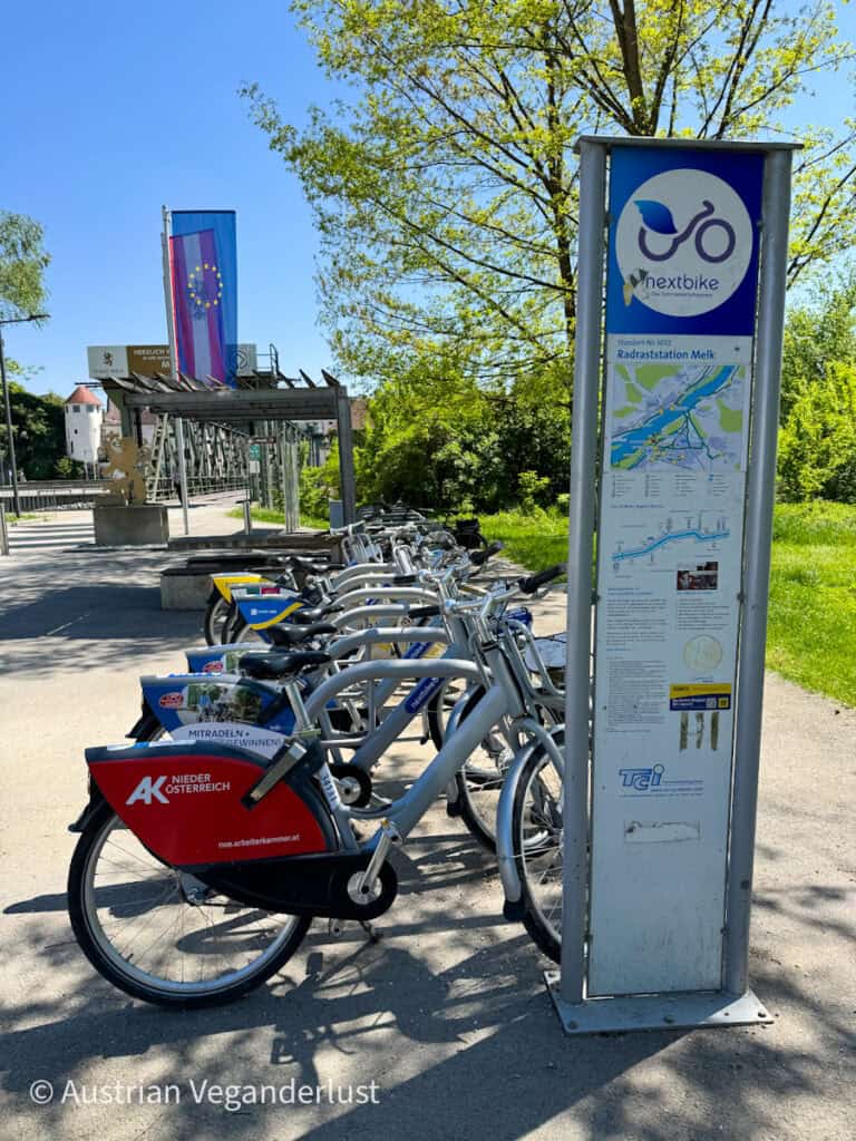 Eine Reihe von silbernen "Nextbike"-Verleihfahrrädern steht an der Radraststation in Melk, Österreich, aufgereiht, mit einem großen Schild, auf dem Karten und Verleihinformationen stehen, umgeben von sonnigem Grün und einer wehenden EU-Flagge.