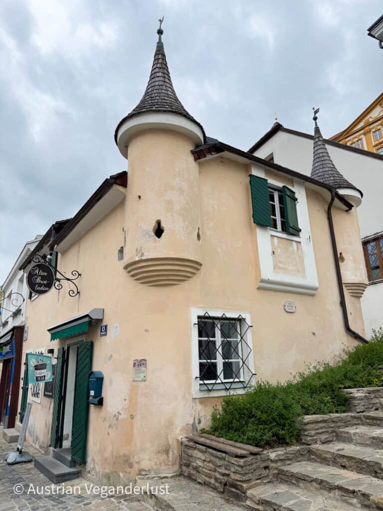 Ein charmantes altes Gebäude in Melk mit kegelförmigen Turmdächern und grünen Fensterläden, in dem sich ein uriger Brotladen mit einem traditionellen Schild befindet.