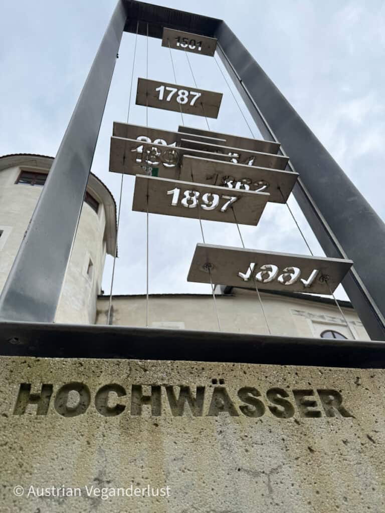 Eine Metallskulptur, die historische Hochwasserstände in Melk, Österreich, anzeigt, mit Jahreszahlen wie 1501, 1787 und 1897, die auf Tafeln über dem in Stein gehauenen Wort "HOCHWÄSSER" hängen.