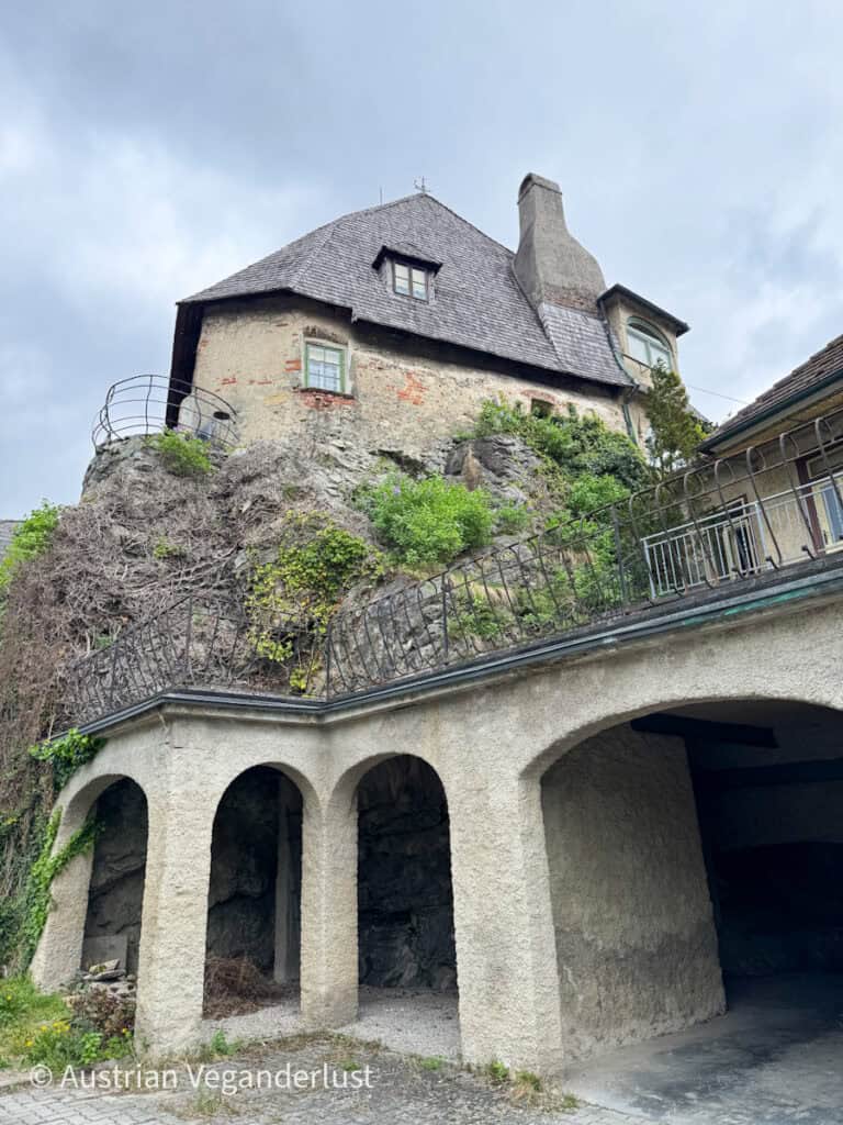 Ein altes Steinhaus, das dramatisch auf einem felsigen Gelände gebaut wurde, mit einer gewölbten Steinstruktur darunter, die sich in die natürliche Umgebung einfügt.