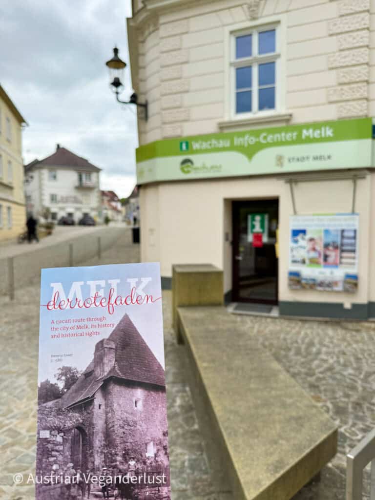 Eine Hand hält eine "Melk der rote Faden"-Broschüre vor dem Wachau Info-Center in Melk, die für einen historischen Wanderweg mit dem alten Brauereiturm wirbt.