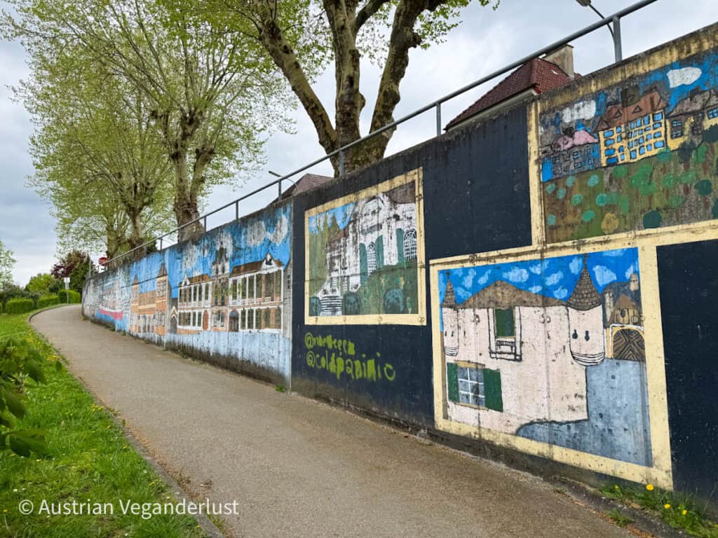 Eine farbenfrohe Wandmalerei entlang eines Spazierwegs in Melk, die lebendige Gemälde von historischen Gebäuden und Wahrzeichen unter Bäumen zeigt.