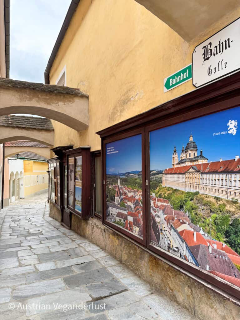 Eine bezaubernde Kopfsteinpflaster-Gasse in Melk, genannt "Bahn-Gasse", gesäumt von gelb gemauerten Gebäuden und großen Fotoplakaten mit Panoramablick auf die Stadt und das Stift Melk.