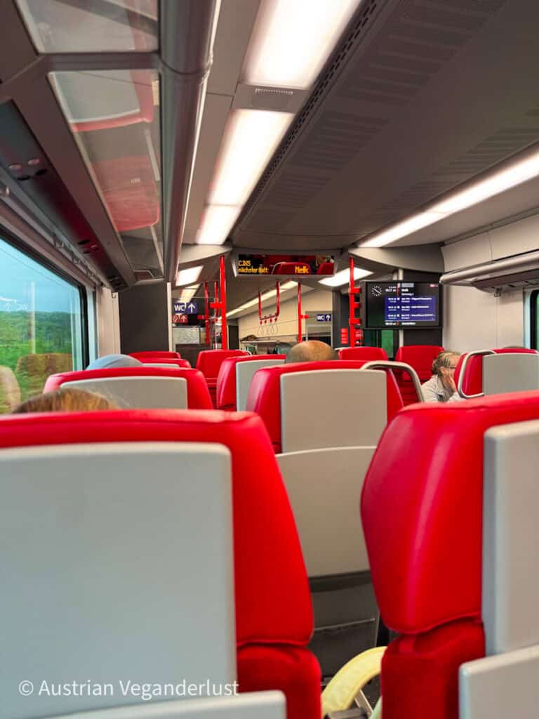 Inside view of a modern Austrian train heading toward Melk, featuring sleek red and gray seats, overhead luggage racks, and electronic displays showing the next stop and other travel information.