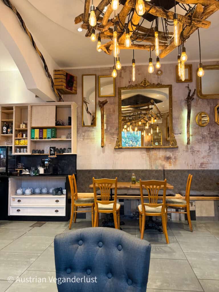 Cozy hostel common room with a rustic wooden chandelier and multiple Edison bulbs hanging from the ceiling. Several gold-framed mirrors decorate the distressed wall, and a light wood shelving unit with drinks and tea sets stands to the left.