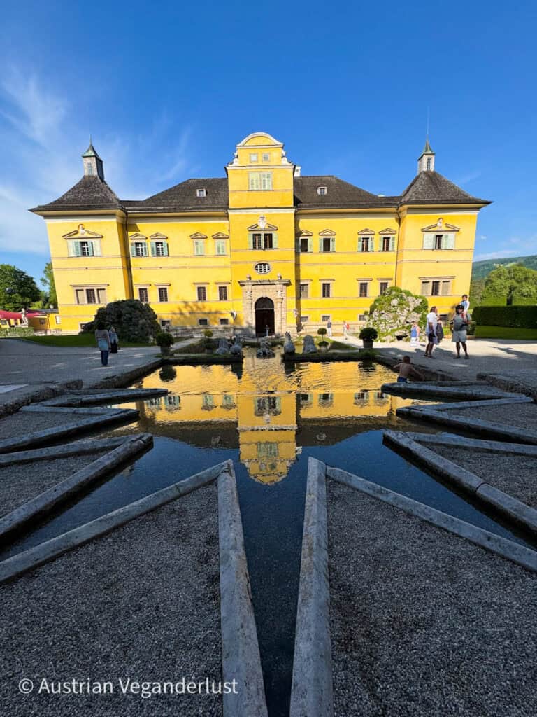 The bright yellow façade of Hellbrunn Palace is perfectly reflected in a decorative fountain pool with stone water channels leading outward. Visitors stroll across the gravel courtyard under a vivid blue sky, enjoying the baroque architecture and tranquil setting.