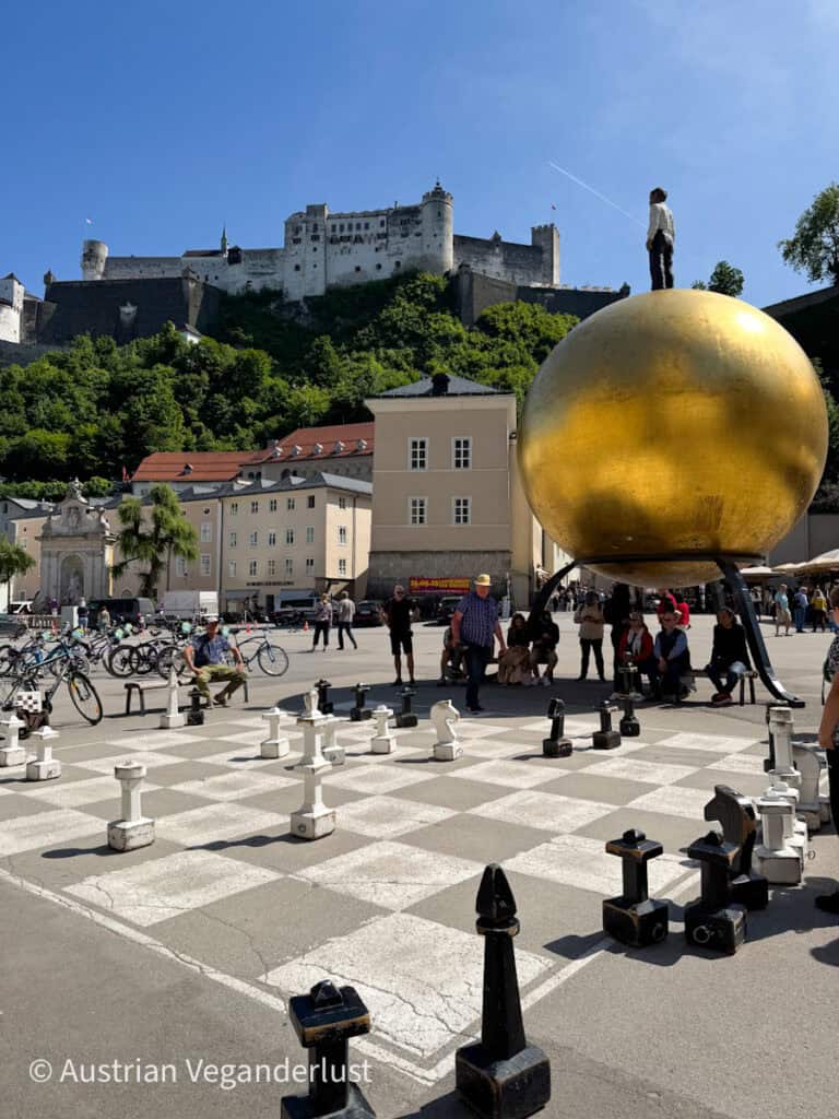 A giant chessboard in Kapitelplatz with people playing in front of the golden Sphaera sculpture, while Hohensalzburg Fortress looms on the hill above.