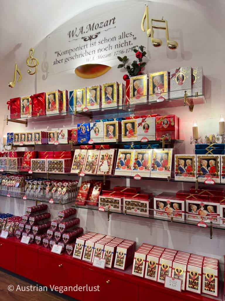 Regale voller bunter Mozart-Schokoladenpackungen und herzförmiger Süßigkeiten mit Porträts von Wolfgang Amadeus Mozart. Goldene Noten zieren die Wand über der Auslage neben dem Text „W.A. Mozart. Komponiert ist schon alles, aber geschrieben noch nicht. W.A. Mozart 1756–1791.“