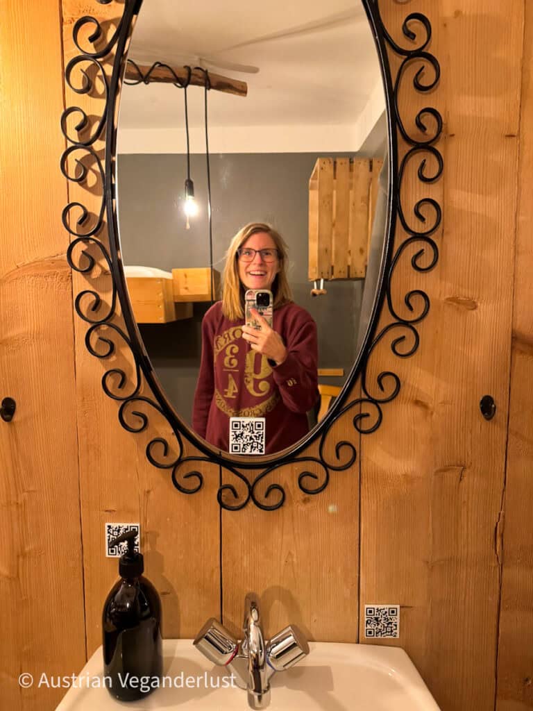 Mirror selfie of a smiling woman with glasses and a burgundy sweater in a rustic hostel room. The oval mirror has an ornate black iron frame, with a washbasin and soap dispenser visible below.