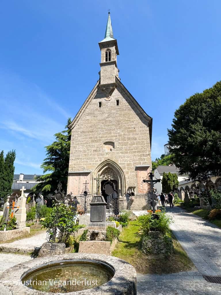 A small stone chapel with a green spire stands in the center of Salzburg’s historic St. Peter's Cemetery, surrounded by ornate wrought iron grave markers and flowering plots. The bright blue sky and cobblestone paths add to the peaceful, picturesque setting.