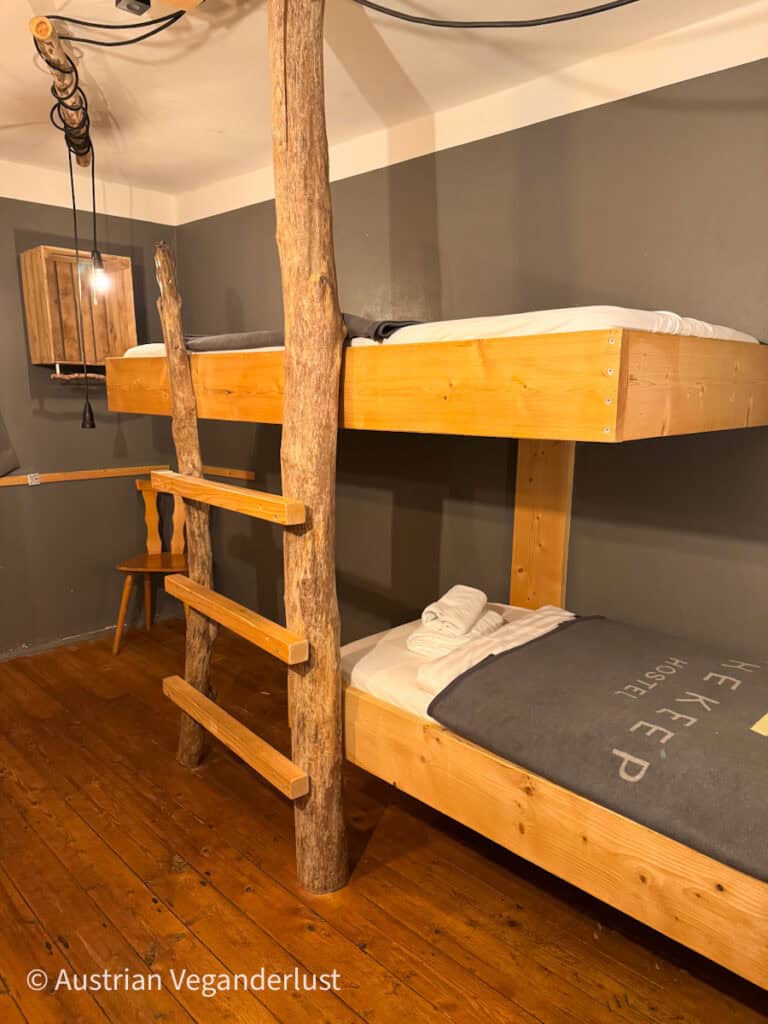 Rustic bunk bed made of light wood with a ladder crafted from real tree trunks in a hostel room. Folded towels are placed on the lower bed, and the bedding features the text "Hostel".