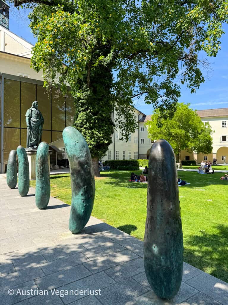 Large bronze sculptures shaped like gherkins stand upright on a walkway in Salzburg, with a statue of a man and leafy park grounds in the background.