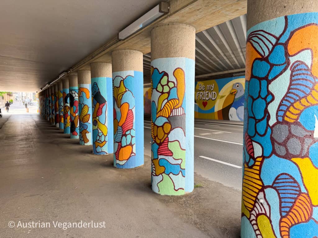 A series of concrete columns under a road overpass in Salzburg are vibrantly painted with abstract, organic street art in shades of blue, orange, red, and yellow. In the background, a cheerful mural on the tunnel wall features a bird and the words “Be my FRIEND” next to a red heart.