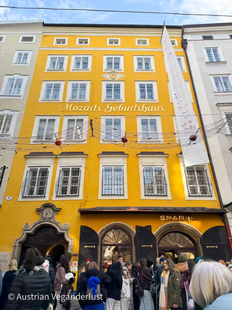 A crowd gathers outside the bright yellow façade of Mozart’s Birthplace (Mozarts Geburtshaus) in Salzburg, Austria. The historic building, now a museum, is marked with ornate white windows, a decorative archway, and a SPAR store on the ground level.