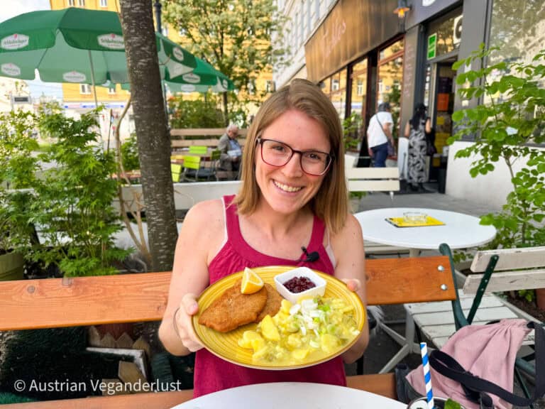 Eine lächelnde Frau in einem Straßencafé hält einen gelben Teller mit einem veganen Schnitzel, einem cremigen Kartoffelsalat mit Frühlingszwiebeln und einer kleinen Schüssel mit Preiselbeersoße in der Hand.