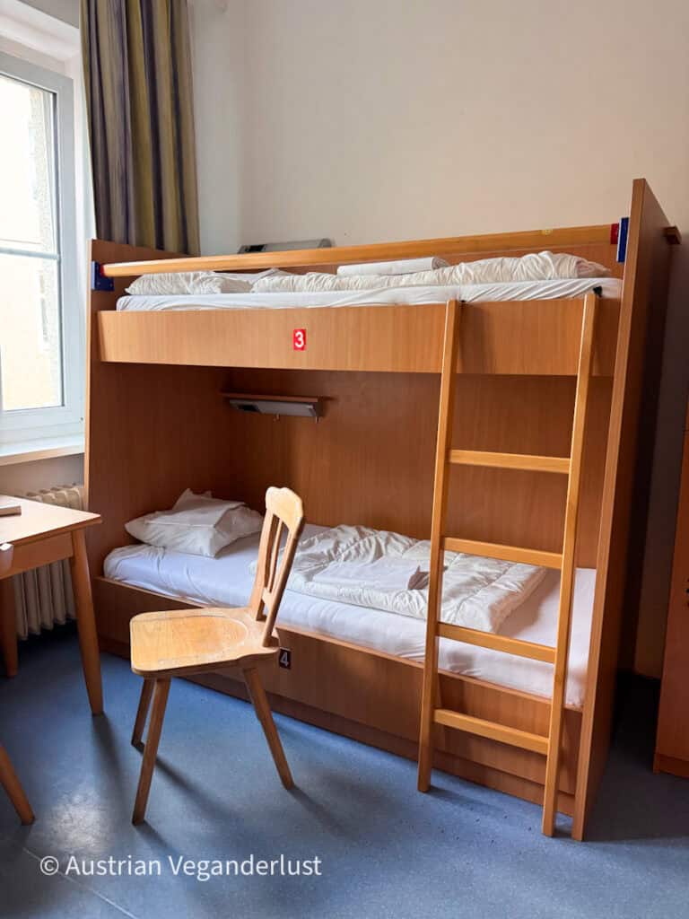 Classic light wood bunk bed with white bedding in a simple hostel room with a blue vinyl floor. The lower bunk is marked with the number "4" and the upper bunk with the number "3".