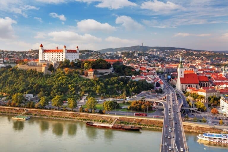 Ein Panoramablick auf Bratislava mit der weiß gemauerten, rot gedeckten Burg von Bratislava, die auf einem Hügel über der Donau thront. Unten führt die SNP-Brücke in die historische Altstadt mit der Martinskathedrale und Reihen von terrakottafarbenen Häusern, die sich in das Tal schmiegen.