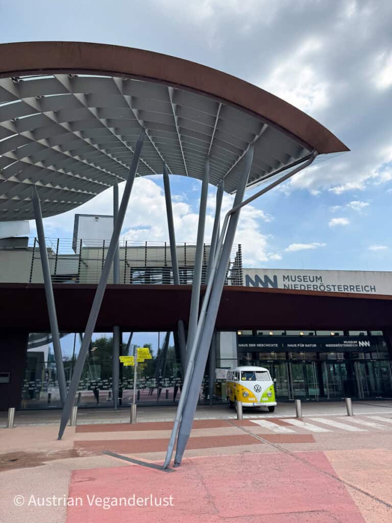 A modern building with an eye-catching curved metal roof and vertical support beams in front of the Museum Niederösterreich. A vintage yellow-and-white VW bus is parked near the glass entrance under a partly cloudy sky.