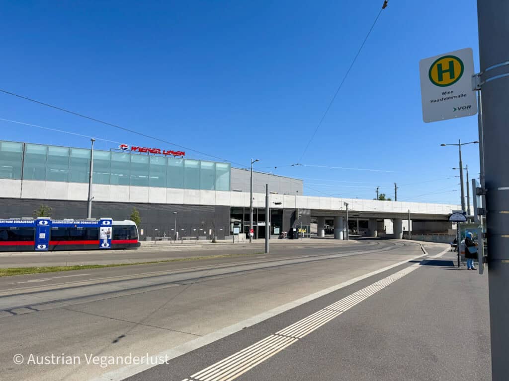 Exterior view of Wien Hausfeldstraße tram station on a clear day, with a red and blue tram waiting and minimal foot traffic.