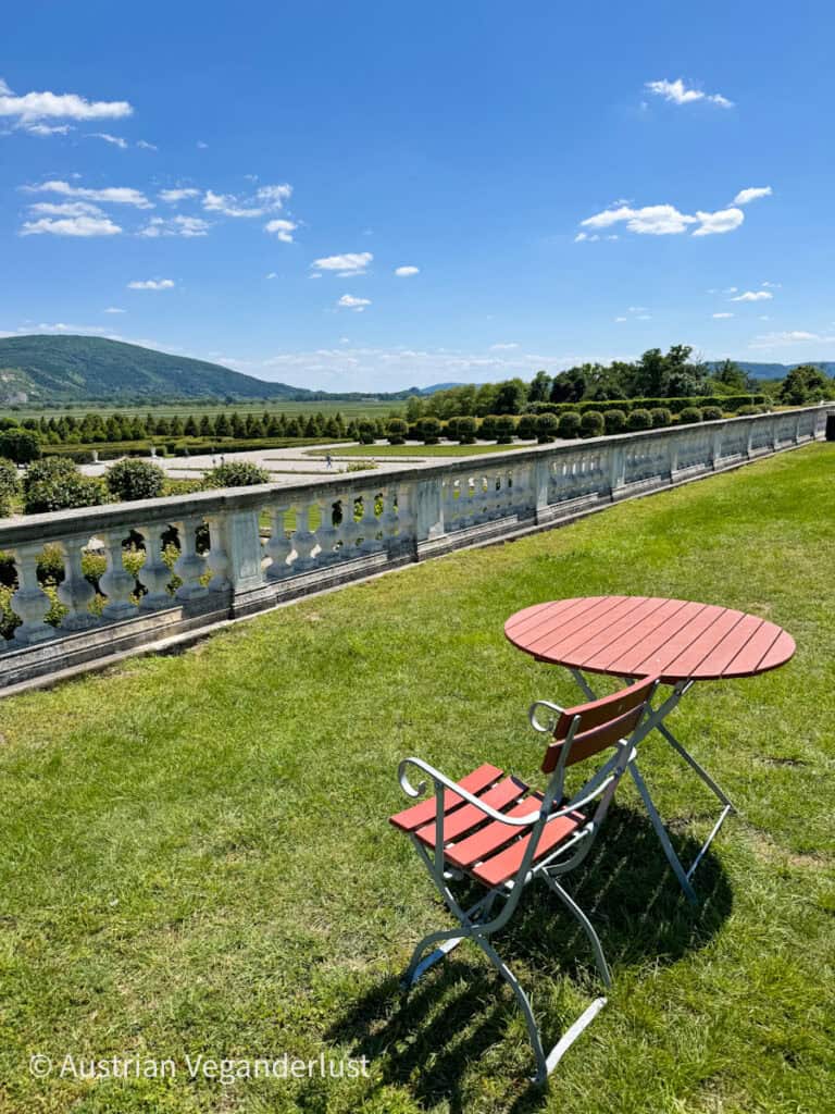 Red metal bistro chair and matching table set on green grass, overlooking the landscaped gardens and distant hills of Schloss Hof.