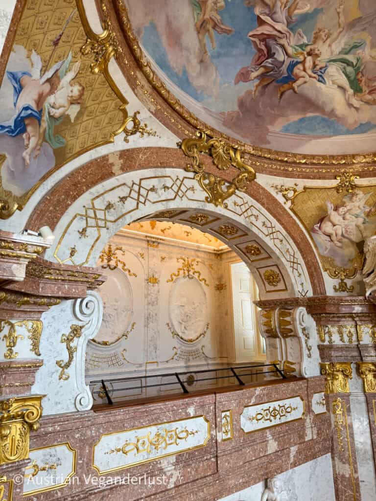Ornate interior view of Schloss Hof, showcasing marble columns, gold accents, and a vibrant ceiling fresco of cherubs and figures in the clouds.