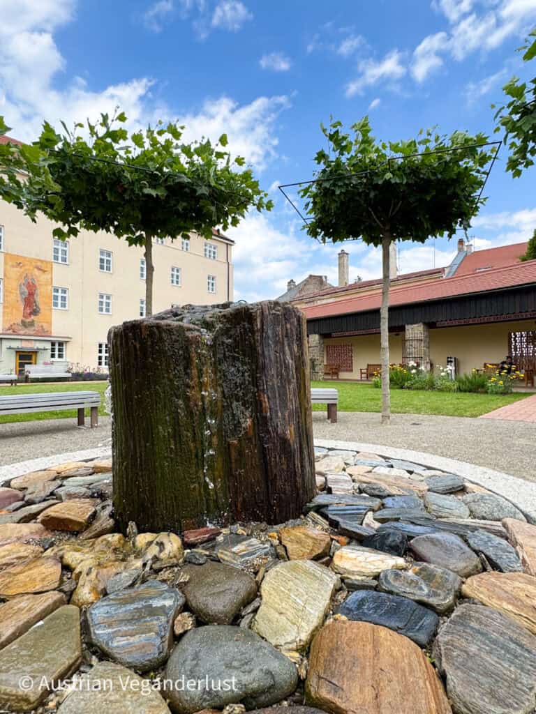 A tranquil garden courtyard with a circular stone fountain made of polished petrified wood, surrounded by neatly placed benches, trimmed trees, and historic buildings in the background.