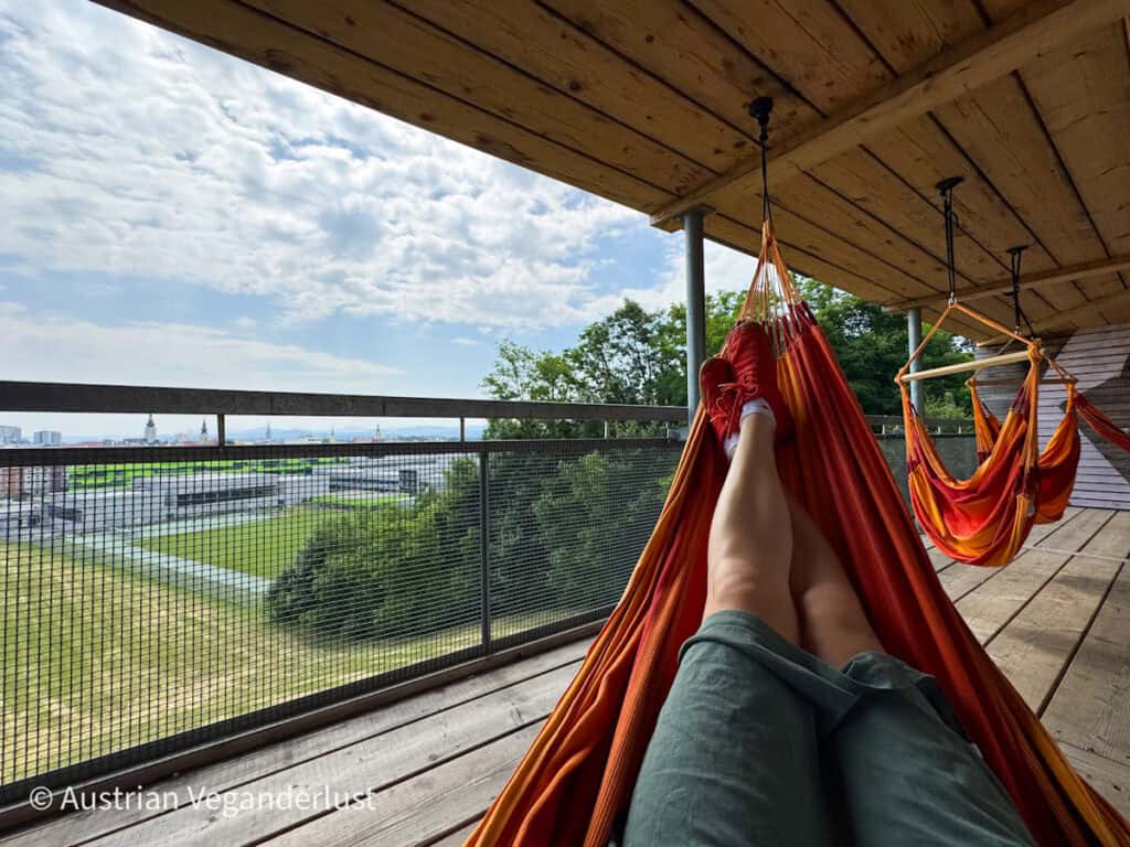 A person relaxes in a red-orange hammock on a wooden balcony at the Flederhaus, overlooking St. Pölten and distant mountains through a mesh safety railing.