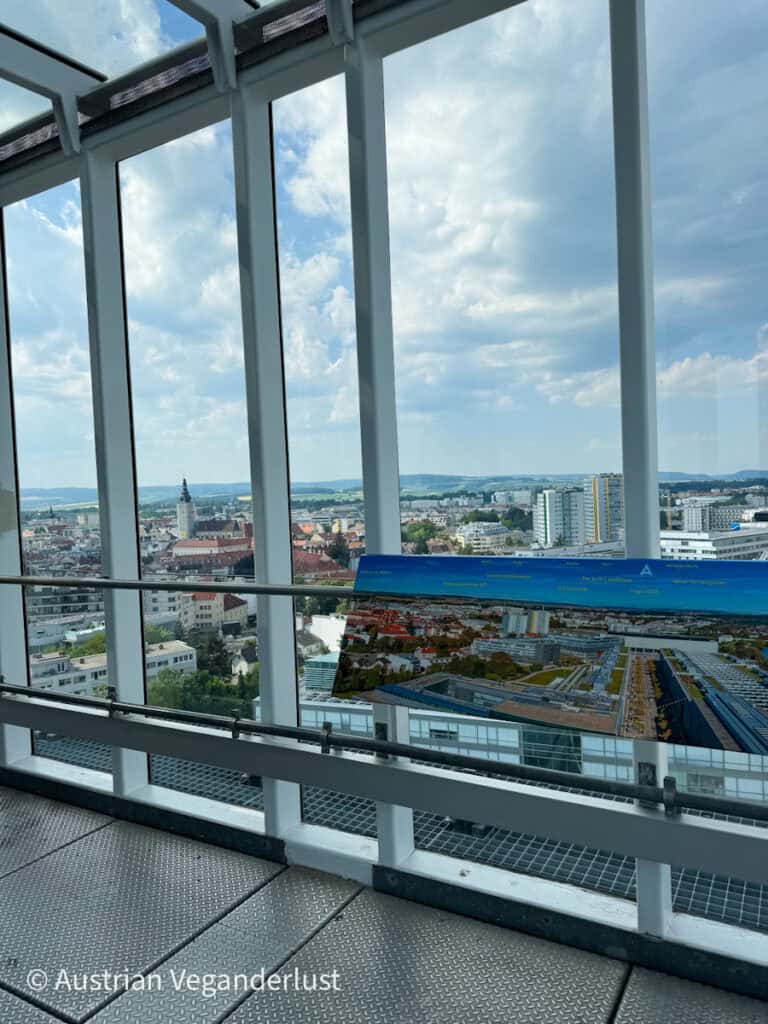 A panoramic view of St. Pölten from the Klangturm observation deck, showing a skyline of church spires and modern buildings through glass windows with a labeled photo guide below.
