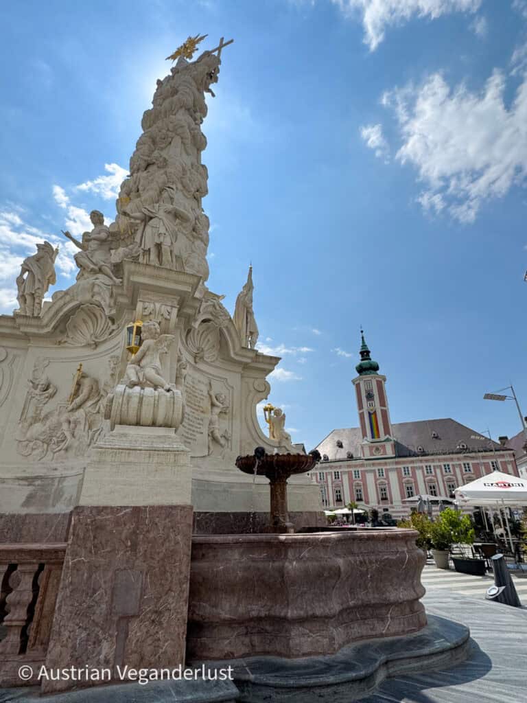 Eine dramatische Aufnahme der verzierten Dreifaltigkeitssäule auf dem Hauptplatz von St. Pölten, mit kunstvollen Statuen und einem Brunnen am Fuß. Das rosa-weiße Rathaus mit der Regenbogenflagge hängt im Hintergrund unter einem strahlenden Himmel.