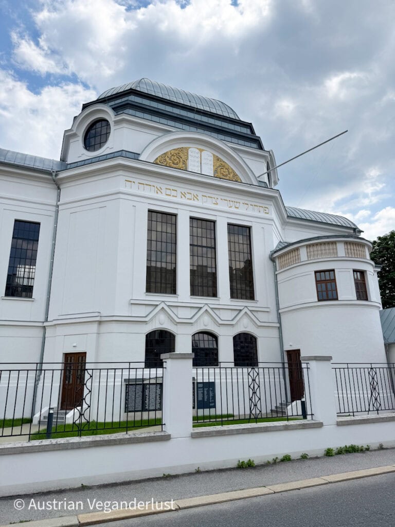 The elegant white façade of the St. Pölten Synagogue, featuring arched windows, Hebrew inscriptions, and a dome-like roof with golden detailing above the entrance.