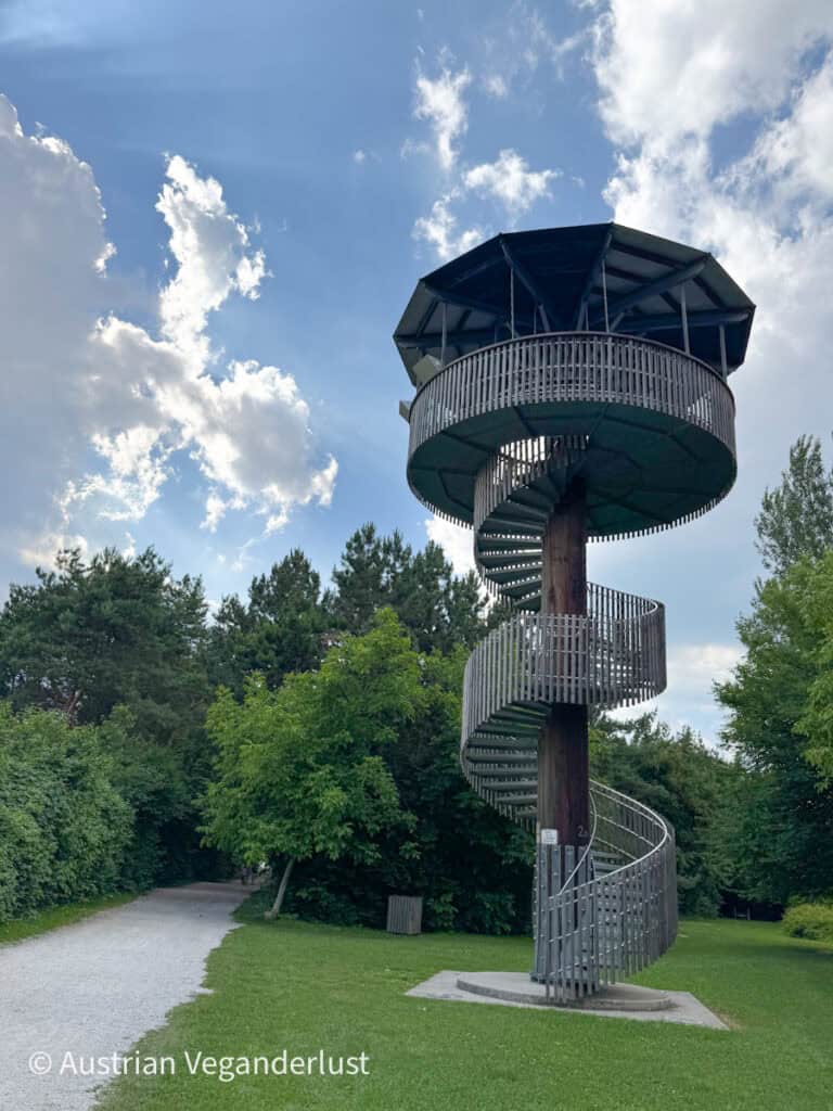 A spiral staircase wraps around a tall wooden observation tower in a lush park area near Viehofner See. The structure is surrounded by trees and set against a bright, cloud-speckled sky.