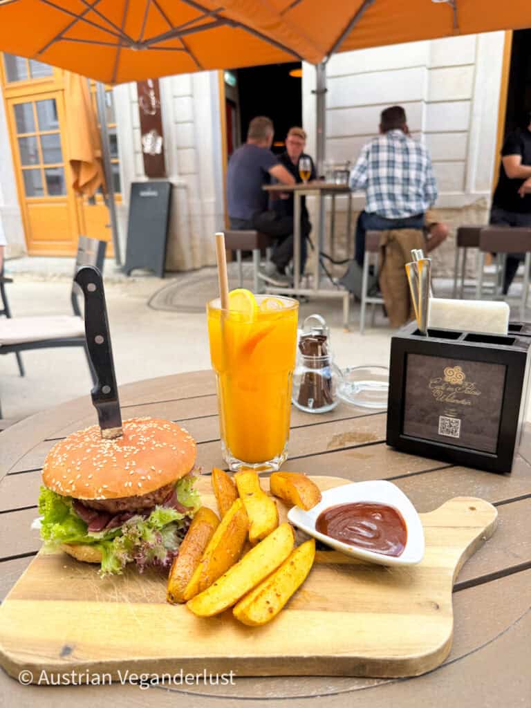 A wooden platter with a vegan burger, potato wedges, and barbecue dip served alongside a glass of orange juice at a café terrace. In the background, people chat at a high table under an orange umbrella.