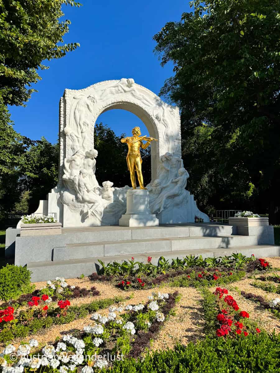 Die goldene Statue von Johann Strauss II., der Geige spielt, steht unter einem mit Skulpturen verzierten Bogen aus weißem Marmor, umrahmt von Bäumen und einem farbenfrohen Blumenbeet im Wiener Stadtpark.