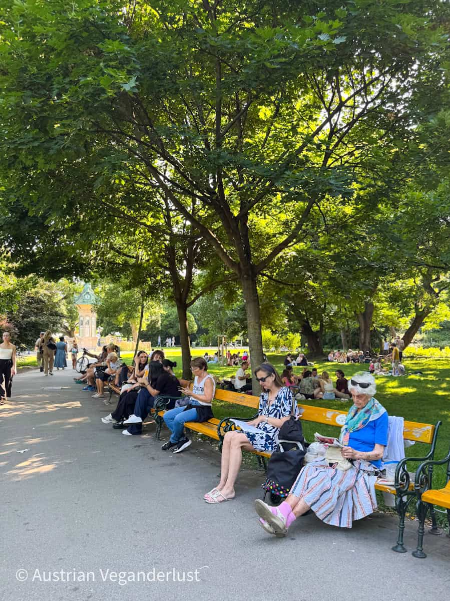 Ein sonniger Tag im Wiener Stadtpark mit Menschen, die sich auf gelben Bänken unter grünen Bäumen entspannen. Im Hintergrund sitzen kleine Gruppen im Gras, während andere den Weg entlang schlendern, auf dem ein kleiner grüner Pavillon steht.