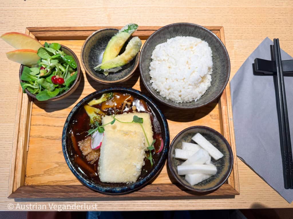 Ein veganes Menü auf japanische Art mit weißem Reis, glasiertem Tofu in einer reichhaltigen Soße, eingelegtem Rettich, frischem Salat mit Apfelscheiben und Tempura-Zucchini auf einem Holztablett.