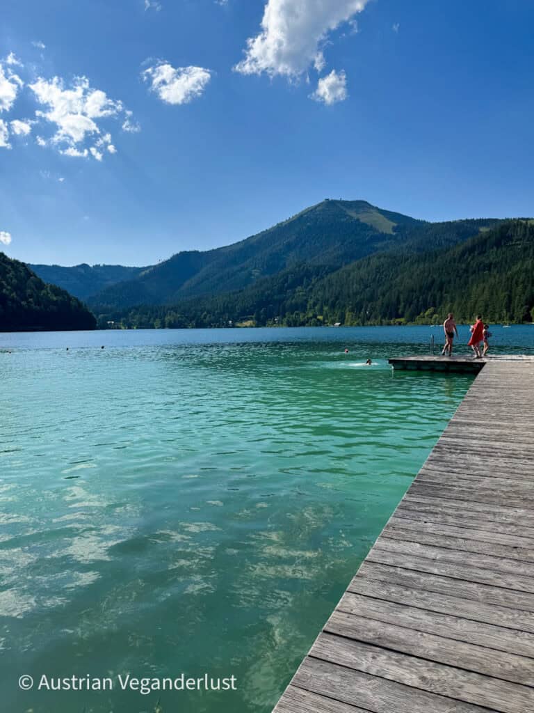 Ein klarer, türkisfarbener Alpensee, umgeben von bewaldeten Bergen unter einem strahlend blauen Himmel, mit einem hölzernen Steg, der ins Wasser ragt, und ein paar Leuten, die die Sonne genießen und schwimmen.