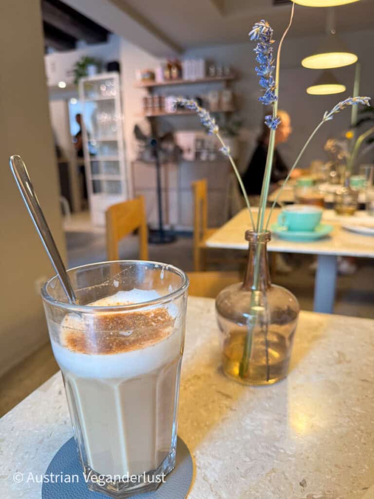 Ein Glas schaumiger Milchkaffee mit Zimtfüllung auf einem Cafétisch mit getrocknetem Lavendel in einer Vase und weicher, gemütlicher Innenbeleuchtung im Hintergrund.