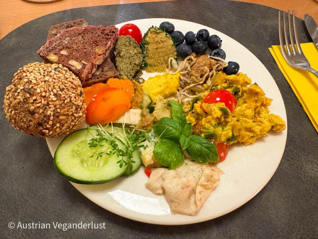 Ein bunter veganer Brunch-Teller mit Körnerbrot, Hummus, Linsenaufstrich, Tofu-Rührei, Kirschtomaten, frischen Kräutern und Früchten wie Heidelbeeren und Aprikosen.